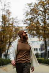 cheerful bearded man in beanie hat and trendy sunglasses looking away against trees and cloudy sky.