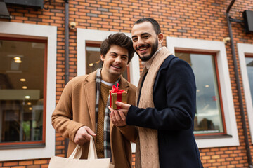 cheerful and stylish gay couple holding shopping bag and Christmas present near blurred store on urban street.