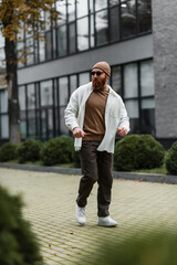 full length of bearded man in beanie hat and stylish sunglasses walking in shirt jacket near modern building.