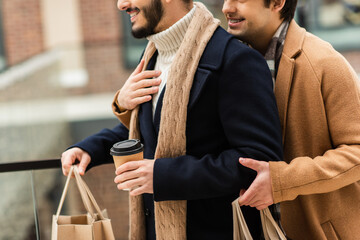 cropped view of smiling gay man hugging bearded boyfriend with coffee to go and shopping bags.