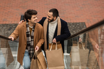 high angle view of gay couple in scarfs holding shopping bags and smiling at each other on...
