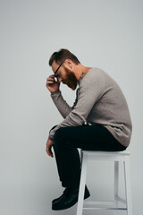 Side view of bearded man touching eyeglasses while sitting on chair isolated on grey.