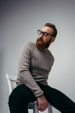 Fashionable bearded man in jumper posing on chair on grey background.