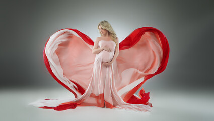 Beautiful pregnant woman in dress with flying fabric that created the shape of a heart. Studio photo, beautiful pose in full growth. Expectant mother gently hugged her tummy. Blonde girl, long hair. © Larysa