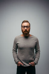 Fototapeta premium Stylish bearded man in jumper holding hands in pockets of pants isolated on grey.