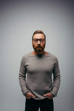 Stylish bearded man in jumper holding hands in pockets of pants isolated on grey.