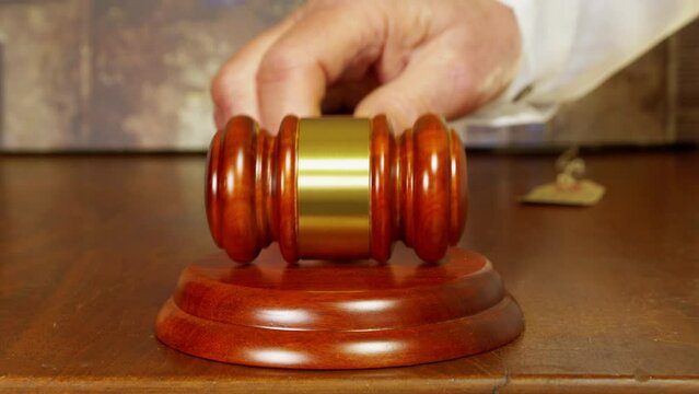 Art auction concept: Closeup of a man&rsquo;s hand striking a traditional wooden gavel against a matching wood block, with part of a defocused painting in the background.