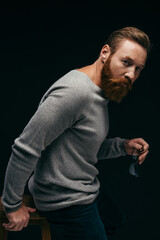 Fashionable man in jumper holding eyeglasses and standing near chair isolated on black.