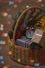 . Wicker basket with gifts and spruce branches. On one of the gifts there is an alarm clock, the arrows of which show 23.55. Wooden background, The photo is decorated with beautiful colored bokeh.


