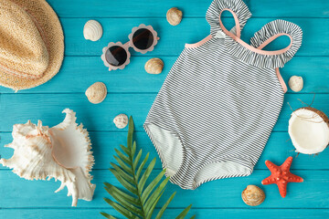 Kids one-piece swimsuit and straw hat next to beach accessories, seashells, starfish and coconut on...