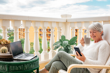 Mature senior woman relaxing on home balcony with a coffee cup while reading message on phone -...