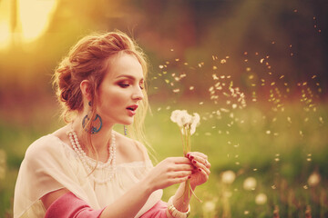 Obraz na płótnie Canvas Beautiful girl in a vintage look sits at sunset in a field of dandelions and blows on dandelions, spring