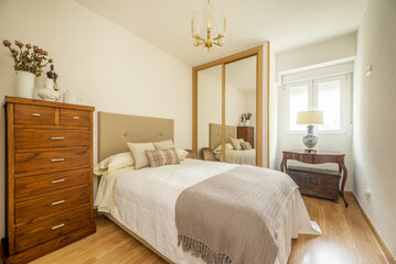 Bedroom with a double bed with a headboard upholstered in beige fabric, a large wooden chest of drawers and a built-in wardrobe on the wall with mirror doors