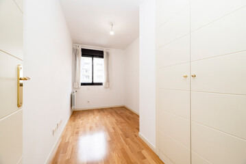 Empty small bedroom with white wooden built-in wardrobe doors, black anodized aluminum windows and white painted walls and wooden floors