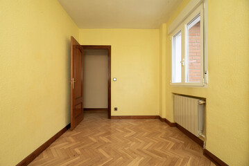 Small empty room with herringbone parquet flooring, niche under window for aluminum radiator and yellow painted walls