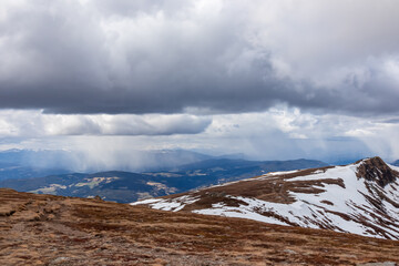 Fototapeta na wymiar Panoramic view on summit cross of mountain peak Gertrusk seen from Ladinger Spitz, Saualpe, Carinthia, Austria, Europe. Snow covered hiking trail over alpine meadows and hills. Cloudy early spring day
