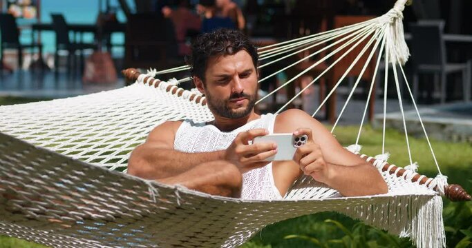Handsome bearded Caucasian young man on vacations, playing virtual online computer games on his smartphone while sunbathing on a hammock. Pleasant guy in summer wear has fun with online entertainments
