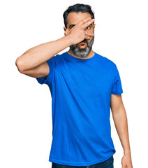 Fototapeta premium Middle aged man with beard wearing casual blue t shirt peeking in shock covering face and eyes with hand, looking through fingers with embarrassed expression.