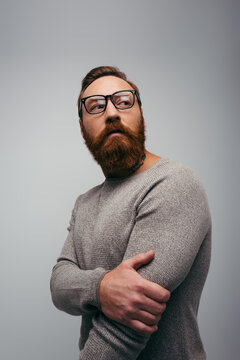 Portrait of stylish bearded man in jumper and eyeglasses looking away isolated on grey.