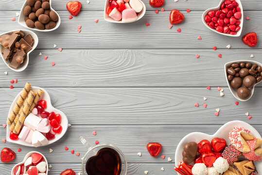 Valentine's Day concept. Top view photo of heart shaped plates with chocolate jelly candies cookies glass of beverage and confetti on grey wooden table background with copyspace in the middle