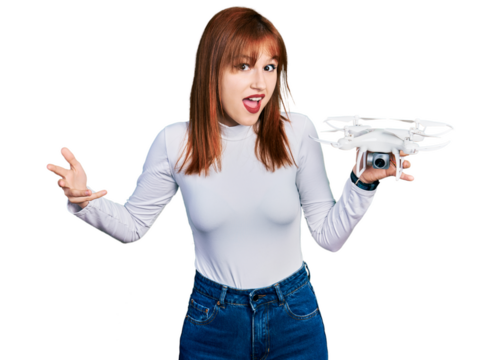 Redhead young woman holding drone celebrating achievement with happy smile and winner expression with raised hand