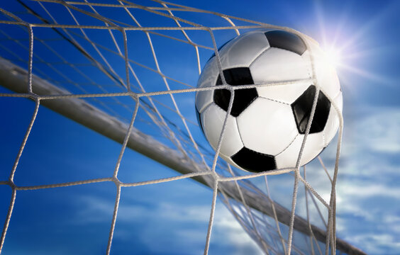 Football or soccer goal, a ball with classic design flying into the net, with blue sky and sun in the background
