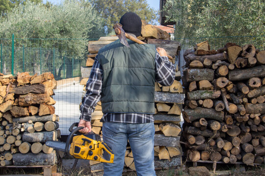 Image of a lumberjack from behind with ax and chainsaw in hand after compiling a pile of wood as preparation for winter. Heating with the home fireplace