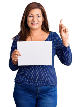Middle age latin woman holding paper banner with blank space surprised with an idea or question pointing finger with happy face, number one