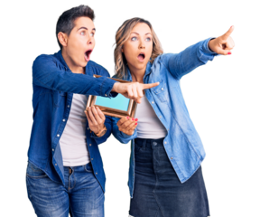 Couple of women holding empty frame pointing with finger surprised ahead, open mouth amazed expression, something on the front