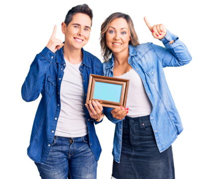 Couple of women holding empty frame smiling amazed and surprised and pointing up with fingers and raised arms.
