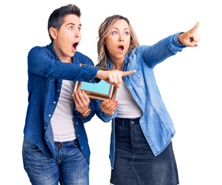 Couple of women holding empty frame pointing with finger surprised ahead, open mouth amazed expression, something on the front