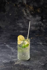 Fotobehang Vertical closeup of a mojito in a clear glass with lemons, lime, and mint on a dark background. © Galip Kürkcü/Wirestock Creators
