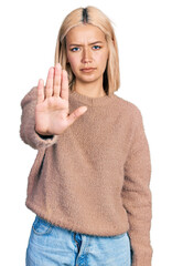 Beautiful young blonde woman wearing brown sweater doing stop sing with palm of the hand. warning expression with negative and serious gesture on the face.