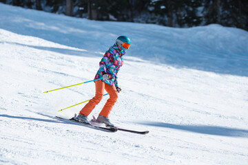 Young skier in bright clothes sliding down the slope on a sunny day at a mountain resort.