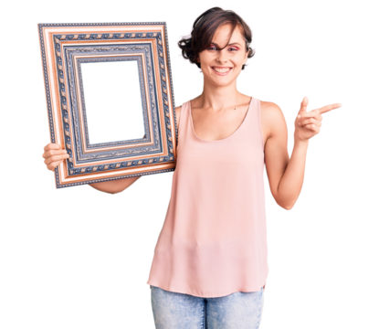Beautiful young woman with short hair holding empty frame smiling happy pointing with hand and finger to the side