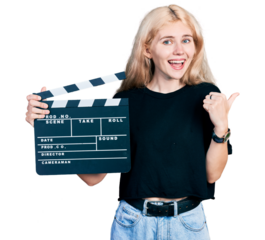 Young caucasian woman holding video film clapboard pointing thumb up to the side smiling happy with open mouth