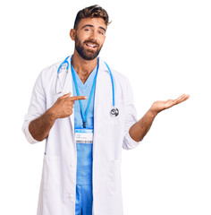 Young hispanic man wearing doctor uniform and stethoscope amazed and smiling to the camera while presenting with hand and pointing with finger.