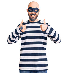 Young handsome man wearing burglar mask success sign doing positive gesture with hand, thumbs up...