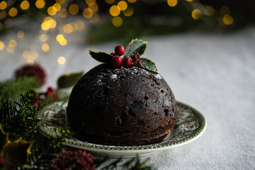 Traditional Christmas pudding decorated with holly on vintage plate, lit candle and natural decorations on linen tablecloth with Christmas lights on background.