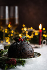 Traditional Christmas pudding decorated with holly on vintage plate, lit candle and natural decorations on linen tablecloth with Christmas lights on background.
