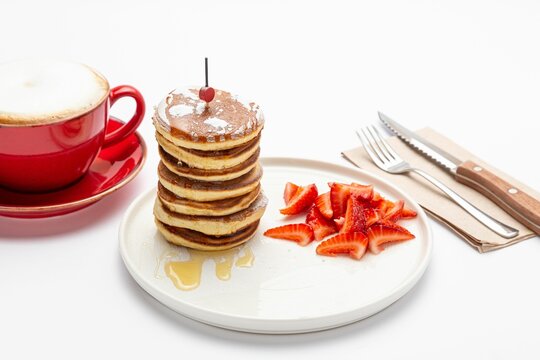 Honey pancakes with a sliced strawberry on a white plate, with a cup of coffee and cutlery