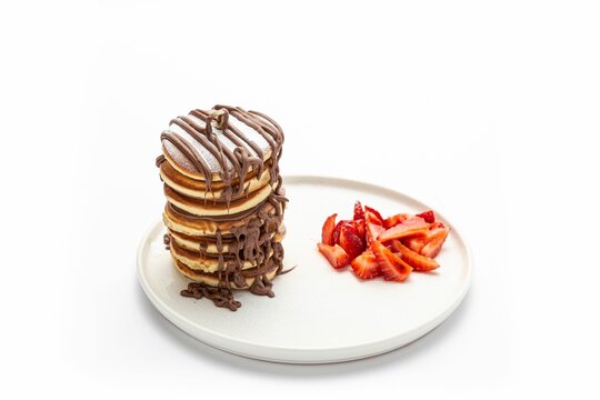 Pile of pancakes covered with chocolate, served with a sliced strawberry on a white ceramic plate