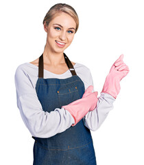 Beautiful young blonde woman wearing cleaner apron and gloves smiling happy pointing with hand and finger to the side