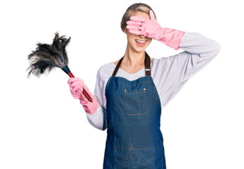 Beautiful young blonde woman wearing apron holding cleaning duster smiling and laughing with hand on face covering eyes for surprise. blind concept.