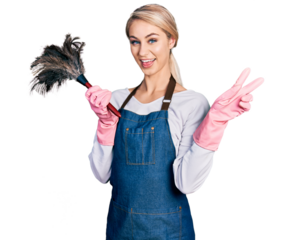 Beautiful young blonde woman wearing apron holding cleaning duster smiling looking to the camera showing fingers doing victory sign. number two.