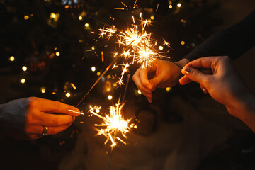 Friends celebrating with burning sparklers in hands against christmas lights in dark room. Happy...