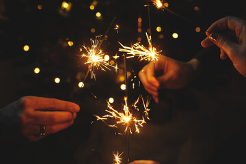 Hands holding fireworks against christmas lights in dark room. Happy New Year! Atmospheric holiday. Friends celebrating with burning sparklers in hands on background of stylish illuminated tree
