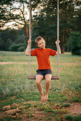 Barefoot boy 7-10 in red t-shirt with bruised legs sitting on rope swing in countryside among pines, in rays of setting sun. Vertical