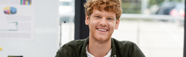 Obraz premium cheerful redhead businessman looking at camera in office, banner
