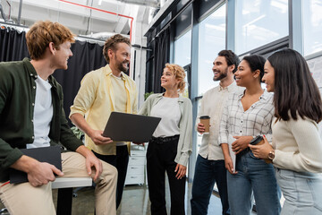 cheerful man with laptop talking to smiling interracial business colleagues in office